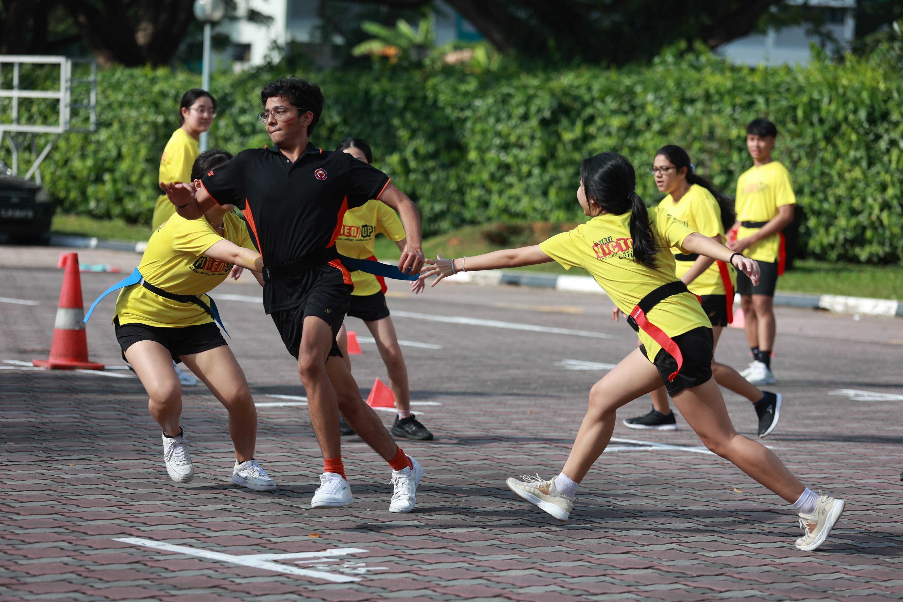 Students in yellow shirts play flag football outdoors, with two players reaching for the flag of a player in black.